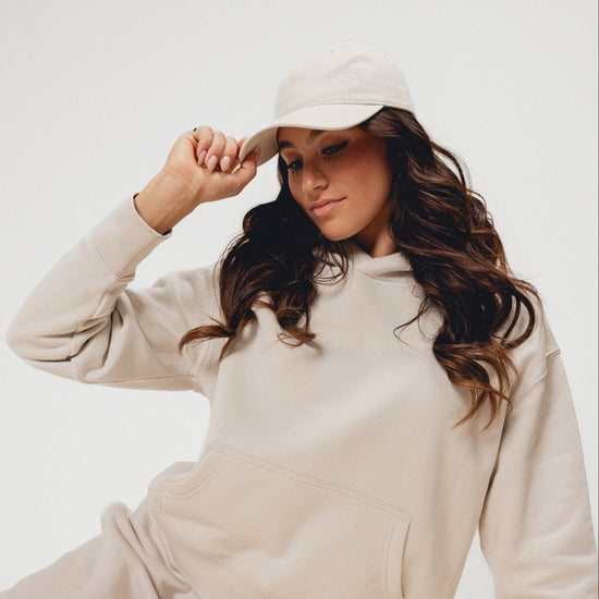 Woman wearing a beige outfit and cap sitting on a white background