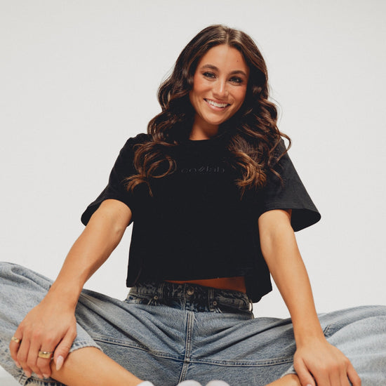 Woman sitting on the floor wearing a black crop top and blue jeans against a plain background