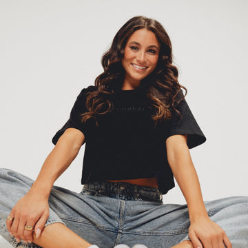 Woman sitting on the floor wearing a black crop top and blue jeans against a plain background