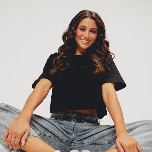 Woman sitting on the floor wearing a black crop top and blue jeans against a plain background