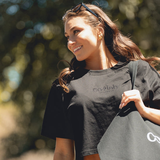 Woman wearing a black t-shirt with colab, holding a matching bag outdoors.