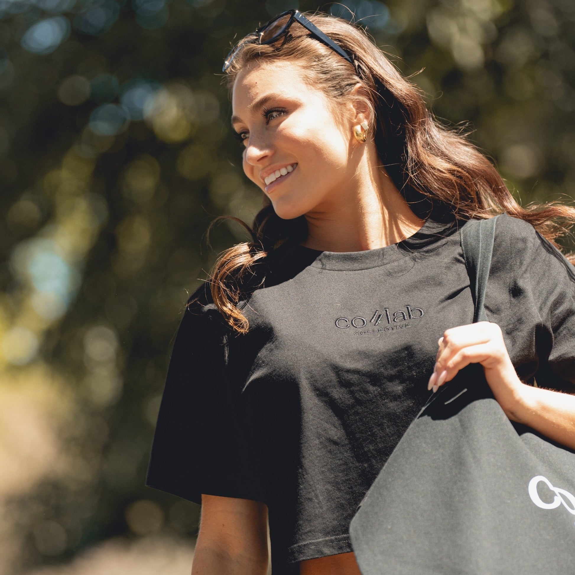 Woman wearing a black t-shirt with colab, holding a matching bag outdoors.